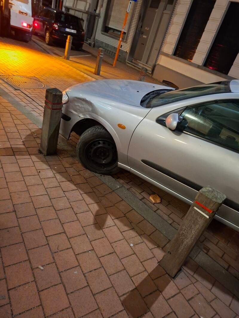 Zilvergrijze wagen met het voorwiel klem tegen een paaltje in een verlichte straat bij nacht